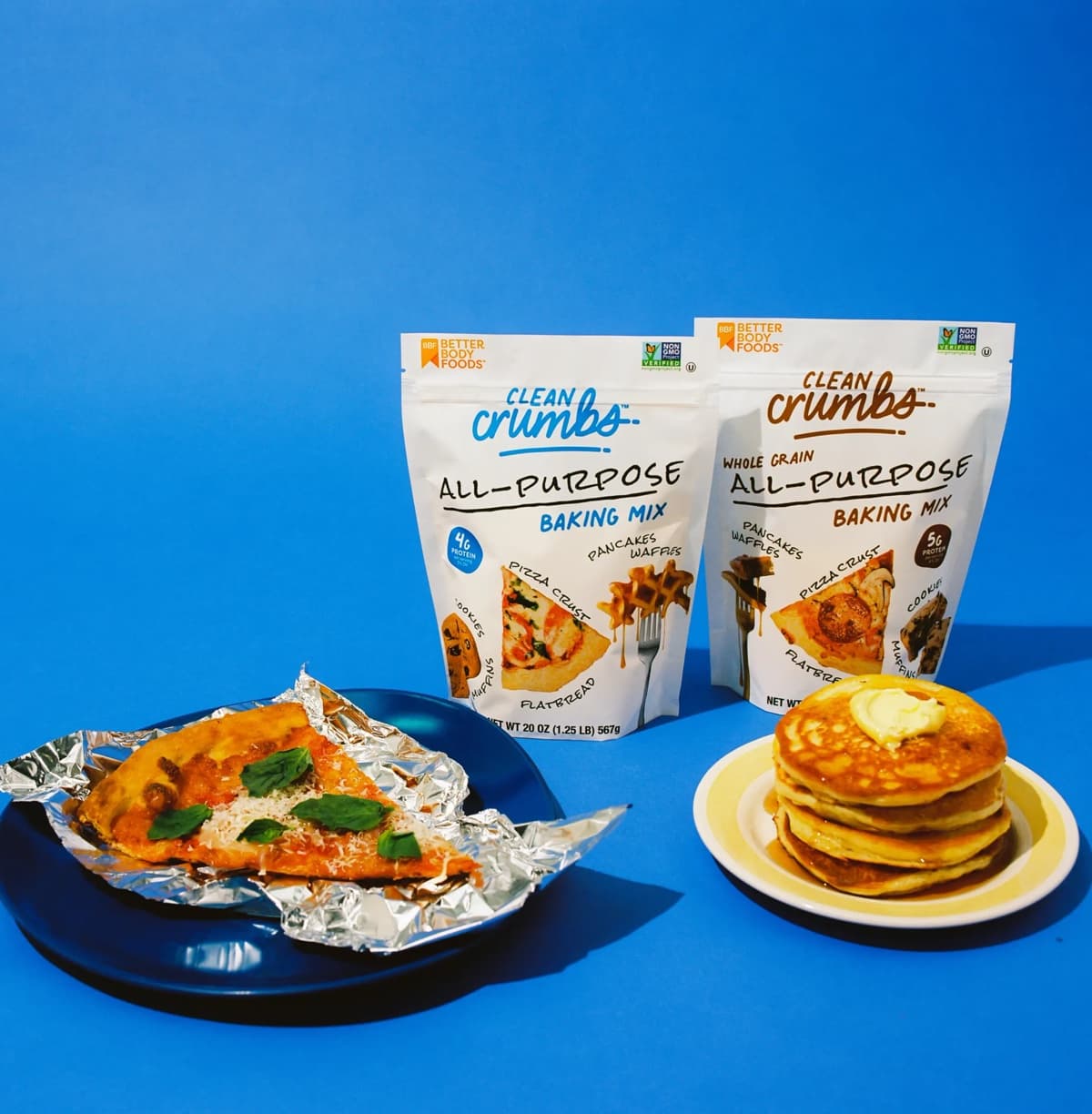 Clean Crumbs baking mixes with a slice of pizza and a stack of pancakes.